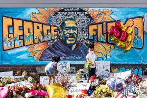 George Floyd mural and memorial. Signs that say "Be the change", and "Black Lives Matter", cover the ground with flower bouquets in between. Two young black children stand in the middle of the pile of signs and flowers and appear to be reading what the signs say.