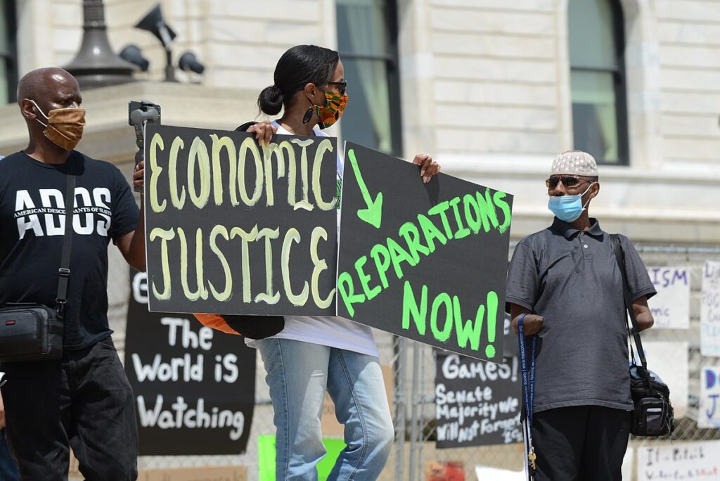 A black woman at a protest holds up two signs that say "Economic Justice" and "Reparations Now". Two black men stand on either side of her.