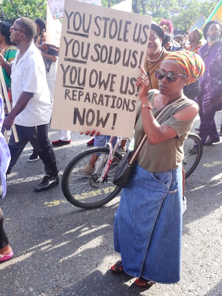 Women on street holding a sign saying You Sold us! You Owe Us! Reparations Now!