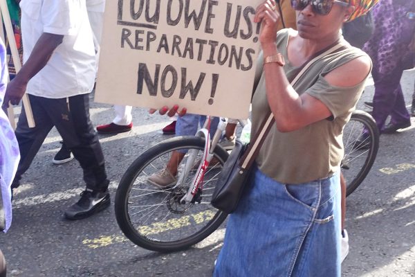 Women on street holding a sign saying You Sold us! You Owe Us! Reparations Now!