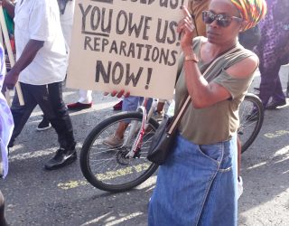 Women on street holding a sign saying You Sold us! You Owe Us! Reparations Now!